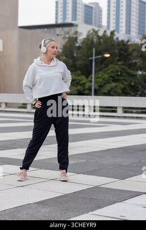 Donna matura che si allena in una piazza pedonale accanto a una ringhiera in cemento con le cuffie Foto Stock