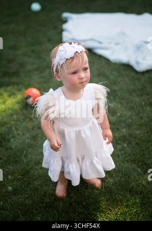 Bella e affascinante ragazza bionda con gli occhi blu, in un abito bianco con una decorazione bianca sulla testa, si erge a piedi nudi sull'erba del giardino. Mostra la demo Foto Stock