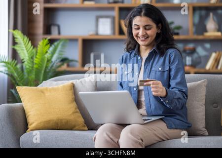 Una giovane donna indiana siede a casa sul divano, tenendo un portatile in grembo e usando una carta di credito in mano. Foto Stock