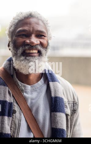 Borsa a tracolla con sciarpa a righe, uomo senior sorridente sul balcone in maglia di denim Foto Stock