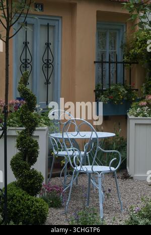 Una pittoresca area salotto presenta delicate sedie blu e un piccolo tavolo, circondato da vibranti fiori e vegetazione in un giardino soleggiato. Foto Stock