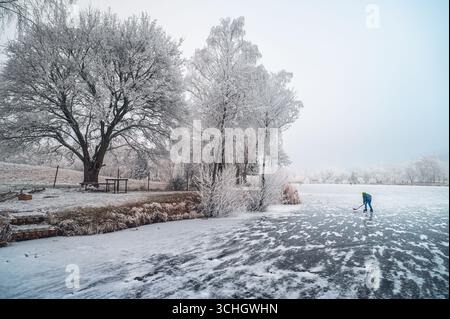 Lo spirito natalizio si anima quando un bambino ama giocare a hockey su uno stagno innevato, circondato da alberi e campi ghiacciati. Foto Stock