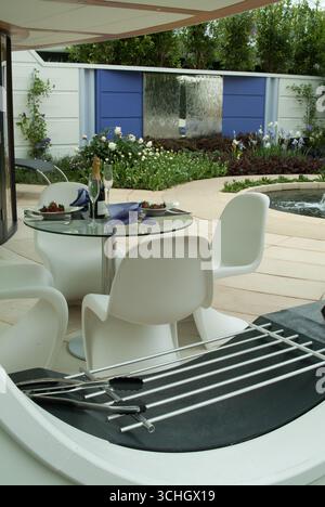 Un'area da pranzo all'aperto presenta eleganti sedie e tavolo, bottiglie di champagne, un barbecue e un vivace giardino sullo sfondo. Foto Stock