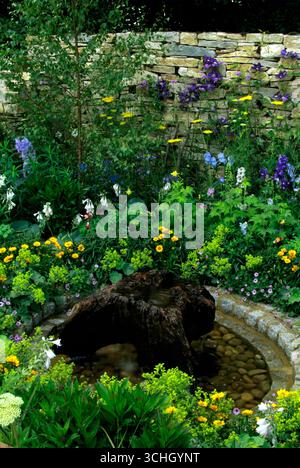 Lussureggiante giardino pieno di una varieta' di fiori luminosi e vegetazione che circondano un tranquillo elemento d'acqua, creando un'atmosfera tranquilla. Foto Stock