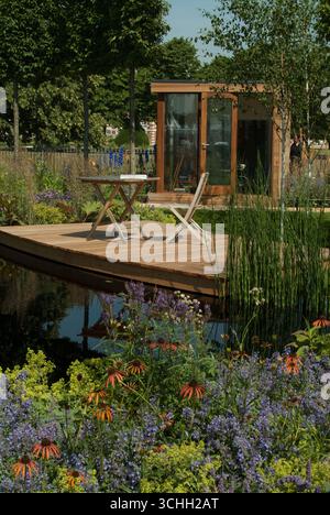 Un tranquillo giardino con un ponte in legno, una cabina moderna e fiori colorati che circondano un tranquillo laghetto in una giornata di sole. Foto Stock