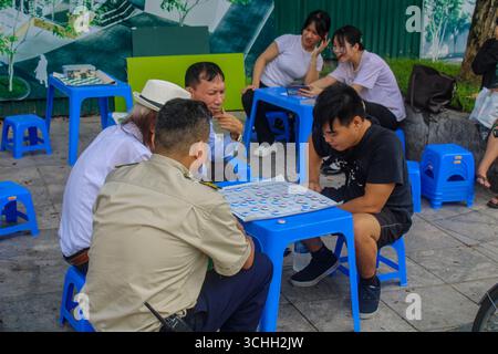Vietnam, Hanoi. Alcune persone stanno giocando a un gioco da tavolo per le strade del centro città. In background, due ragazze si divertono a guardare i video su Foto Stock