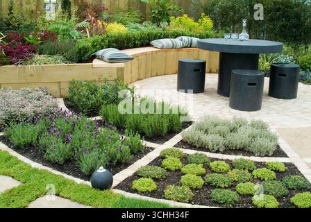 L'invitante giardino presenta un tavolo contemporaneo e un'area salotto circondata da piante vibranti ed erbe fresche in un ambiente tranquillo. Foto Stock