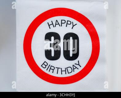 Un vibrante segno circolare rosso mostra 'Happy 30th Birthday' in caratteri bianchi. Il cartello è posizionato su una superficie bianca, probabilmente all'interno di un ristorante o ce Foto Stock