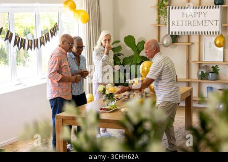 Festeggiamo il pensionamento, amici anziani diversi che bevono bevande e spuntini in una camera decorata, a casa Foto Stock