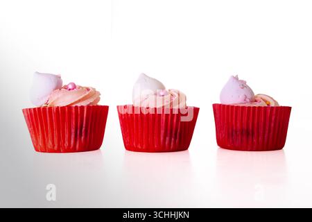 Tre cupcake rosa con glassa a vortice, topper di meringa e perle di zucchero in involucri rossi, disposti fianco a fianco su sfondo bianco. Foto Stock