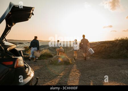 Vista posteriore dei membri della famiglia che camminano verso grandi rocce sulla riva del mare per un picnic Foto Stock