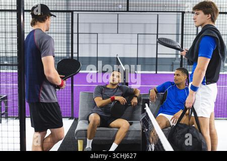 Diversi amici maschi che riposano sul divano chiacchierando sul campo del padel con racchette e borsa con asciugamano Foto Stock