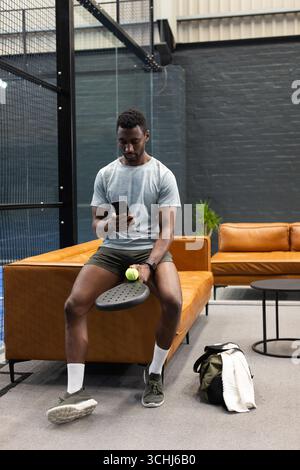 Uomo afro-americano in abbigliamento sportivo seduto nella palestra con lo smartphone paddle check Foto Stock