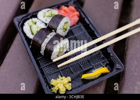 Deliziosi sushi roll ripieni di cetriolo sono disposti ordinatamente in un vassoio nero insieme a wasabi, zenzero sottaceto e una fetta di limone in un picnic rustico Foto Stock