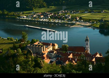 Soleggiata vista ad alto angolo della storica Dürnstein con tetti rossi, il Danubio con una chiatta di passaggio, un campeggio sulla riva opposta e una piccola h Foto Stock