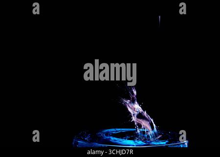 La goccia d'acqua di colore blu colpisce il bordo di vetro, generando uno spruzzo diagonale con increspature su uno sfondo scuro, catturato con tecnica macro ad alta velocità. Foto Stock