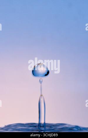 La goccia trasparente si libra sopra una sottile colonna di rimbalzo su una superficie calma su uno sfondo blu pallido, scattata con la fotografia macro ad alta velocità a gocce d'acqua. Foto Stock