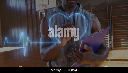 Osservando il fitness coach in felpa con cappuccio che monitora il battito cardiaco sul campo della palestra con fischio e appunti Foto Stock