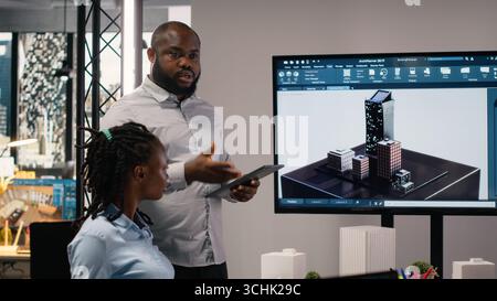 Ingegnere immobiliare afro-americano che presenta il piano di preparazione del sito e le normative sulla suddivisione in zone sui modelli 3D durante una sessione di collaborazione con il team. Iniziativa abitativa per la pianificazione urbana. Foto Stock
