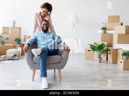Felice sorridente giovane afro americano marito seduto in poltrona, moglie in piedi accanto a lui in appartamento con scatole di cartone Foto Stock