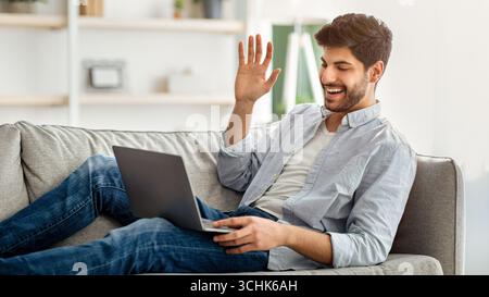 Concetto di comunicazione a distanza. Emozionato uomo arabo videochiamata su computer portatile, sdraiato su divano, spazio libero Foto Stock