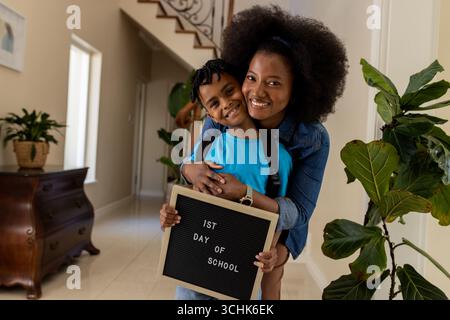 Madre e figlio sorridono, tenendo in mano un cartello per il primo giorno di scuola a casa Foto Stock