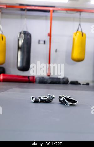 Guanti da combattimento in bianco e nero appoggiati su un tappetino imbottito in palestra, con sacchetti da pugno appesi Foto Stock