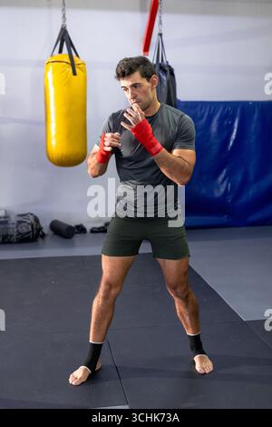 Pugile maschile in piedi in posizione boxe sul tappeto in palestra con sacchetto di punzonatura giallo Foto Stock