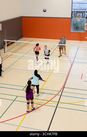 Diverse squadre femminili di abbigliamento sportivo giocano a pallavolo su rete nella palestra scolastica con panchine e cerchio Foto Stock