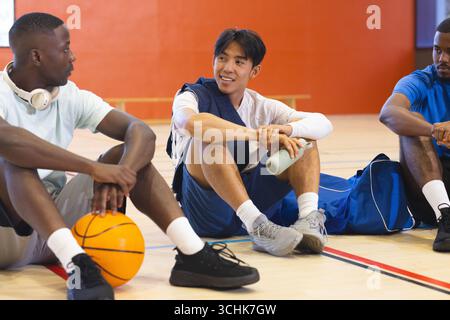 Diversi partner maschili che riposano e chiacchierano sul campo da palestra tenendo in mano il borsone con bottiglia d'acqua da basket Foto Stock