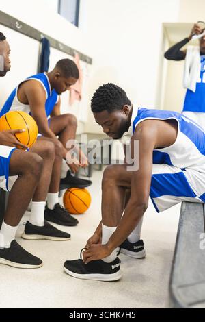 Partner afroamericani maschi che si preparano in spogliatoio legando scarpe sulla panchina con palle di palle Foto Stock