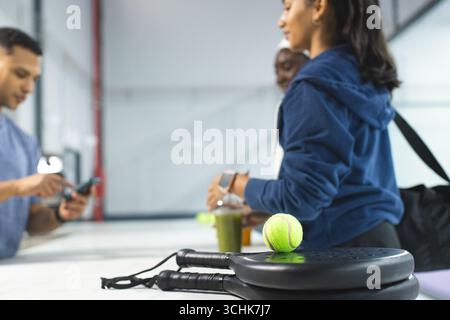 Amici diversi che utilizzano smartphone e carte e aspettano alla reception con racchette da tennis Foto Stock