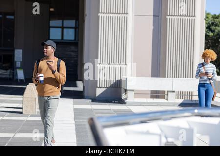 Uomo afroamericano che cammina in piazza indossando auricolari, tenendo in mano una tazza di caffè, uno zaino, uno spazio per le copie Foto Stock
