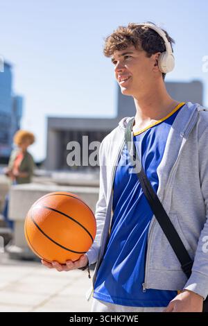 Uomo che si trova in piazza e tiene in mano il pallacanestro con cuffie con cappuccio, cinturino per borsa e spazio per copiare Foto Stock