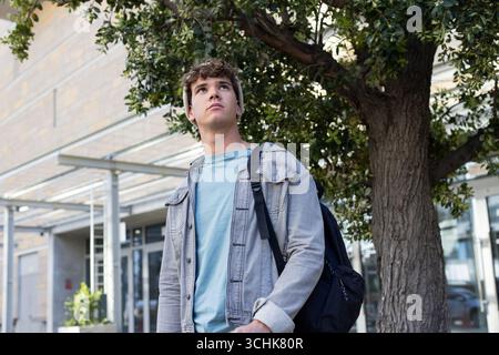 Studente maschio che fa una pausa sotto l'albero davanti a un ingresso moderno che indossa un berretto in maglia, portando con sé uno zaino scuro Foto Stock