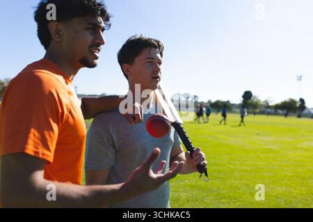 Diversi compagni di squadra maschi discutono della tecnica di battitura sul campo di erba con mazza e palla da cricket Foto Stock