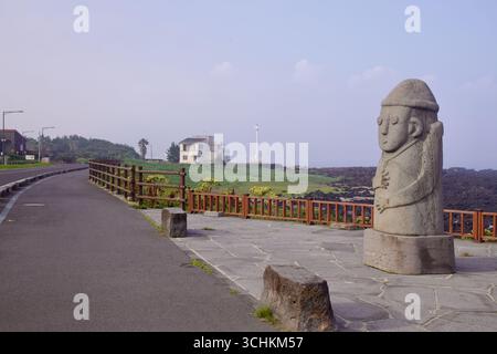 Una classica statua in pietra di Jeju dolhareubang si affaccia sulla costa di Sagye, accanto a una recinzione in legno e a una passeggiata lastricata, con un faro basso sbiadito sul Foto Stock