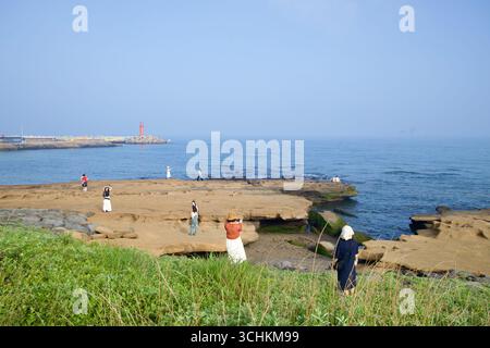 I visitatori camminano attraverso ampie distese di tufo tagliate dalle onde sulla costa di Sagye, con piscine di marea e alghe verdi lungo i bordi e un lontano faro rosso Foto Stock