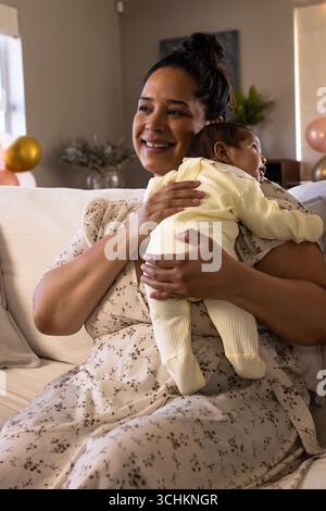 Madre sorridente che tiene il neonato sul divano durante la festa di famiglia a casa Foto Stock