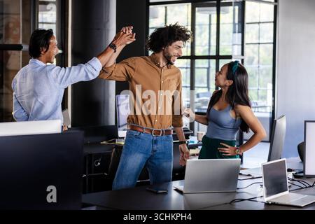 Team diversificato che celebra il successo in un ufficio moderno con laptop sulla scrivania Foto Stock