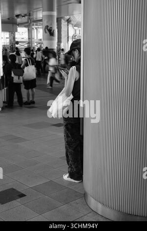 Una giovane donna si trova dietro un pilastro nel centro di transito urbano, assorbita dal suo smartphone, catturando solitudine, concentrazione e ruolo della tecnologia nella navigazione mod Foto Stock