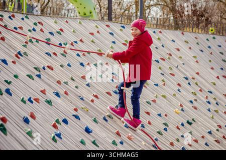Bambino felice che si arrampica su una parete di roccia artificiale usando la corda sul parco giochi Foto Stock