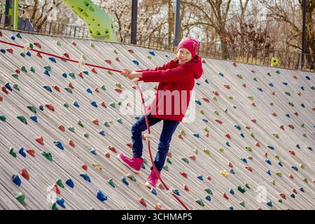 Bambino felice che si arrampica su una parete di roccia artificiale usando la corda sul parco giochi Foto Stock