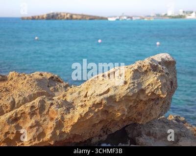Vista da dietro di una grande roccia porosa gialla su uno splendido mare Mediterraneo blu, la spiaggia di Nissi, una piccola isola e gli edifici alberghieri sullo sfondo. Foto Stock