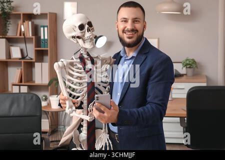 Giovane uomo d'affari con scheletro umano e telefono cellulare in ufficio Foto Stock