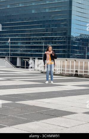 Donna in piazza vicino alla ringhiera esterna dell'edificio in vetro che indossa una sciarpa rosa che trasporta una borsa marrone Foto Stock