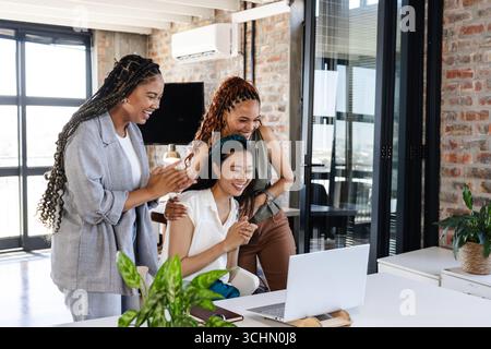 Diverse colleghe che celebrano il successo, riunite attorno a un notebook in un moderno ufficio Foto Stock