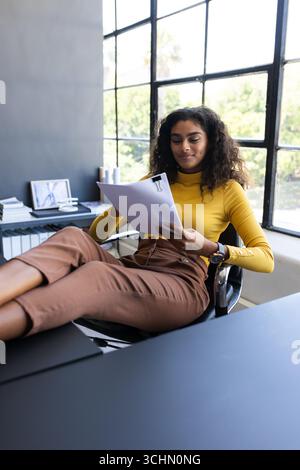 Donna sicura di sé in maglione giallo che esamina documenti con i piedi sulla scrivania, in ufficio e in ufficio Foto Stock