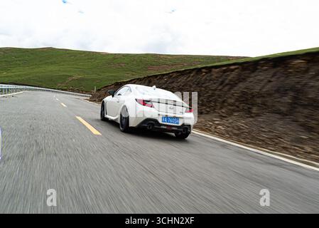 Una ripresa dinamica del tracciamento dell'angolo posteriore di una coupé sportiva bianca che accelera lungo una tortuosa autostrada di montagna delimitata da pendii verdi. Foto Stock