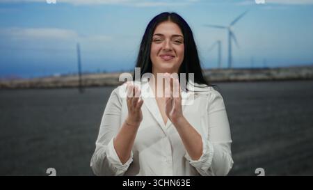 Donna in piedi in un campo ventoso con mulini a vento sullo sfondo che battono, indossando una camicia bianca, sorridendo con fiducia e celebrando la bellezza di Re Foto Stock
